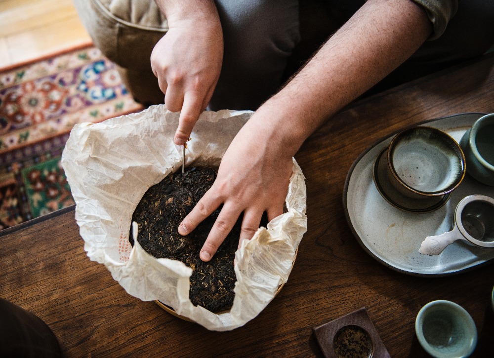 Person breaking a pressed tea cake with a knife on a wooden table, next to teacups.