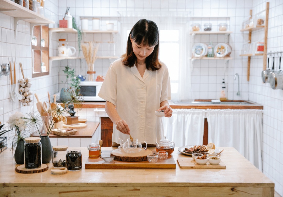 Woman preparing tea in a cozy kitchen with jars and snacks on the counter.