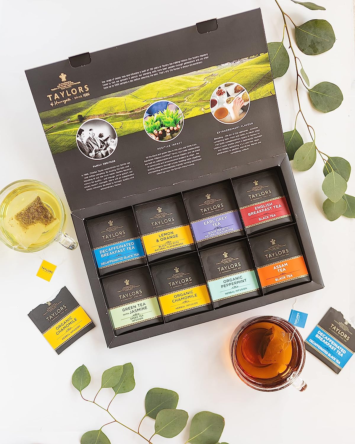 Open box of Taylors tea variety pack with two cups of tea and eucalyptus leaves.